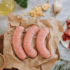 Overview shot of three uncooked sausages on a brown paper on a wooden board. Ingredients surround it, including olive oil, a pot of dried herbs, garlic cloves, some crushed garlic and some cut up beef fillet with some sprigs of thyme.
