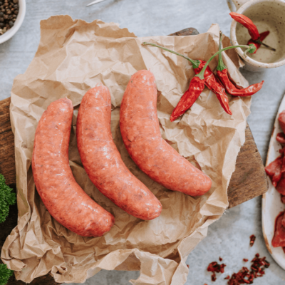Overview shot of three uncooked sausages on a brown paper on a wooden board. Ingredients surround it, including a pot of black peppercorns and some dried red chillies and cut up beef fillet with parsley.