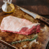 Waipawa Butchery Beef Tri Tip sits on a tray with a small glass bottle of olive oil behind a sprig of rosemary in front and a small butchery knife next to it.