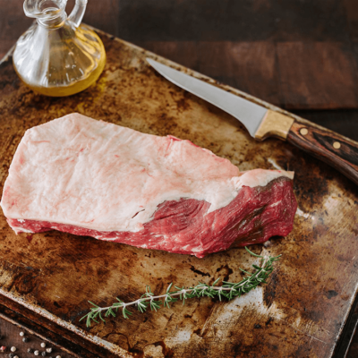 Waipawa Butchery Beef Tri Tip sits on a tray with a small glass bottle of olive oil behind a sprig of rosemary in front and a small butchery knife next to it.