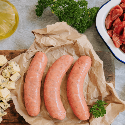Overview shot of three uncooked sausages on a brown paper on a wooden board. Ingredients surround it, including olive oil, cubed blue cheese, a pot of peppercorns and some cut up beef fillet with parsley.