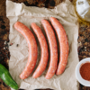 Four uncooked Lamb Merguez sausages sit on some paper on a roasting pan, bottom left is a chilli, bottom right is a small round white dish filled with paprika and a small glass bottle of olive oil sits at the top right of screen.