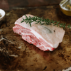 A lamb rump sits on top of a tray with a sprig of rosemary on top with a bowl of peppercorns and olive oil sitting behind.