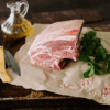 A lamb shoulder sits on brown paper next to sprigs of fresh mint with a small glass bottle of olive oil behind and a knife next to it.