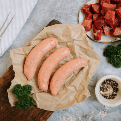 3 uncooked Waipawa Beef Sausages sits on some brown paper on a board with parsley next to it. A plate a chopped beef sits behind and a small white bowl of peppercorns sits in front.