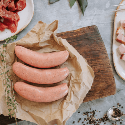 Overview of three uncooked sausages laying on brown paper on a wooden board. The ingredients surround them, includig cut up bacon, cut up beef fillet, and black peppercorns.