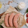 Overview shot of three uncooked sausages on a brown paper on a wooden board. Ingredients surround it, including olive oil, a pot of horseradish, pink salt and peppercorns and some cut up beef fillet with parsley.