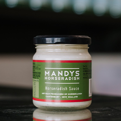 A jar of Mandys Horseradish sits on a reflective bench with out of focus bottles behind it