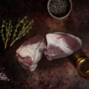 Two uncooked lamb hearts sit on a chopping board with some thyme springs, oil and pepper
