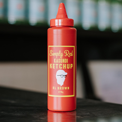 A bottle of Al Brown's Kasundi Ketchup sits on a reflective bench with some out of focus bottles in the background.