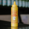 A bottle of Al Brown's old yella habanero mustard sits on a reflective bench with some out of focus bottles in the background.