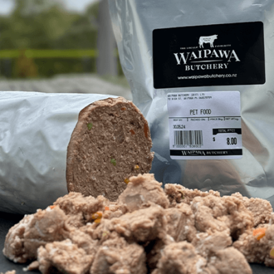 Cut up pet food roll sits on the table with a pouch of dog food sitting behind. Out of focus greenery is in the background.