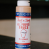 A bottle of Al Brown turf 'n' surf all purpose sauce sits on a reflective bench with some out of focus bottles in the background.